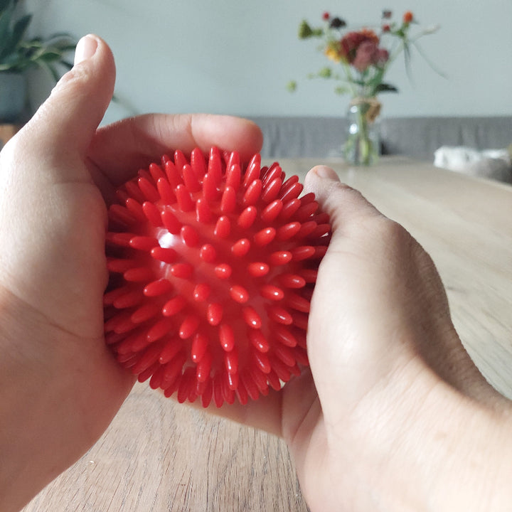roter massageball in händen, an einem Holztisch mit blumen im hintergrond, sensorischer massage ball, 