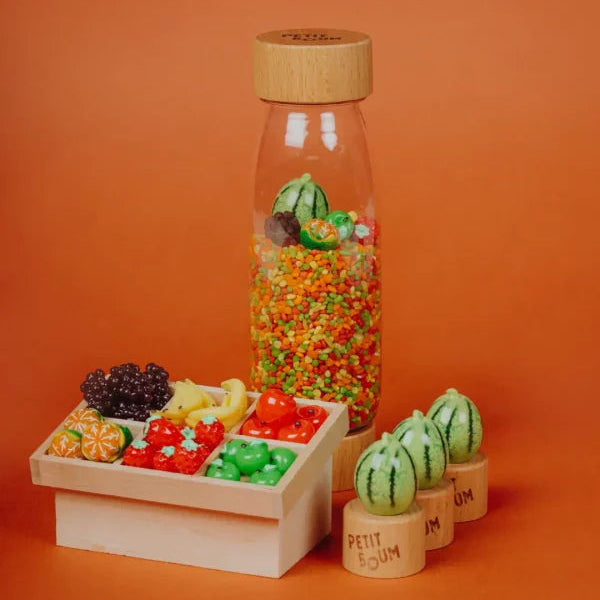 foto met oranje achtergrond, sensoriek fles van petit boum met fruit en confetti stukjes in groen, oranje, rood en geel, op de voorgrond fruitkiste met het fruit dat ook in de fles zit, si materiaal